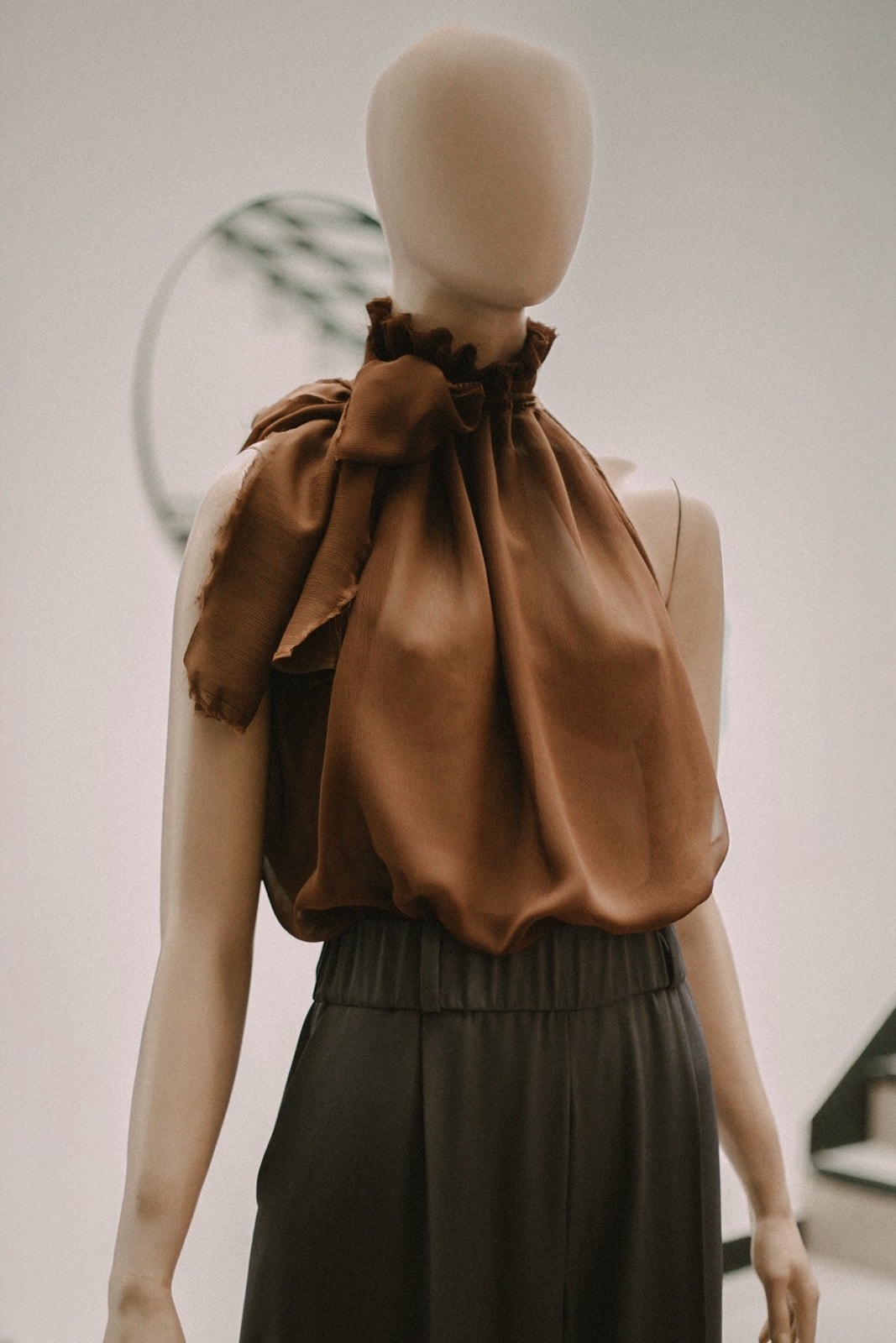Detalle de perfil del cuello alto de la blusa de gasa chocolate con el lazo anudado sobre el hombro, sobre maniquí.
