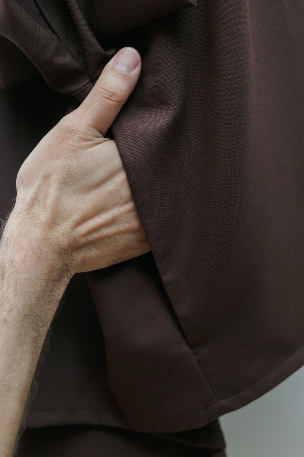 Detalle de la tela de casimir marrón de la campera, con la mano rozando el pliegue interior para mostrar la caída del tejido.