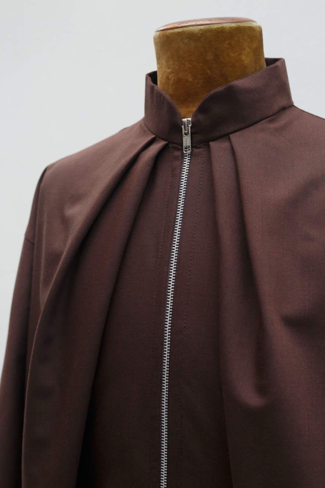 Detalle del cuello alto y los pliegues frontales de la campera de casimir marrón, con el cierre metálico plateado en primer plano, sobre maniquí.