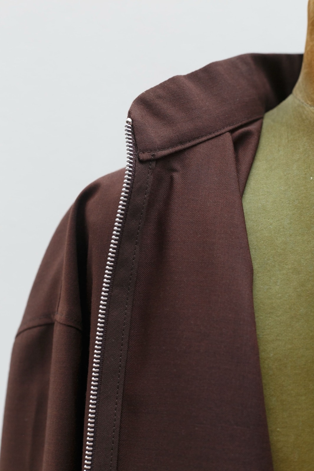 Detalle del hombro y la costura del cuello alto con el cierre metálico plateado de la campera marrón, sobre fondo claro.
