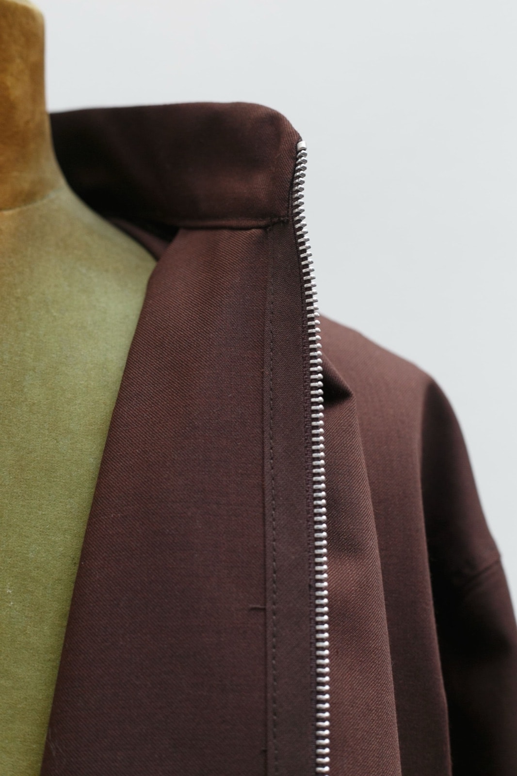 Primer plano del cuello alto y el nacimiento del cierre metálico junto al hombro de la campera marrón, sobre maniquí y fondo claro.