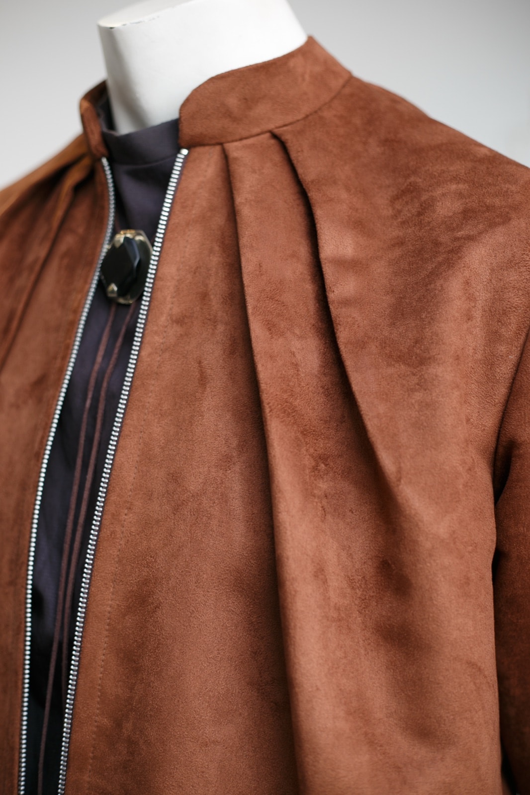 Detalle del cuello alto y los pliegues frontales de la chaqueta de gamuza caramelo, con un broche oscuro visible sobre la prenda interior.