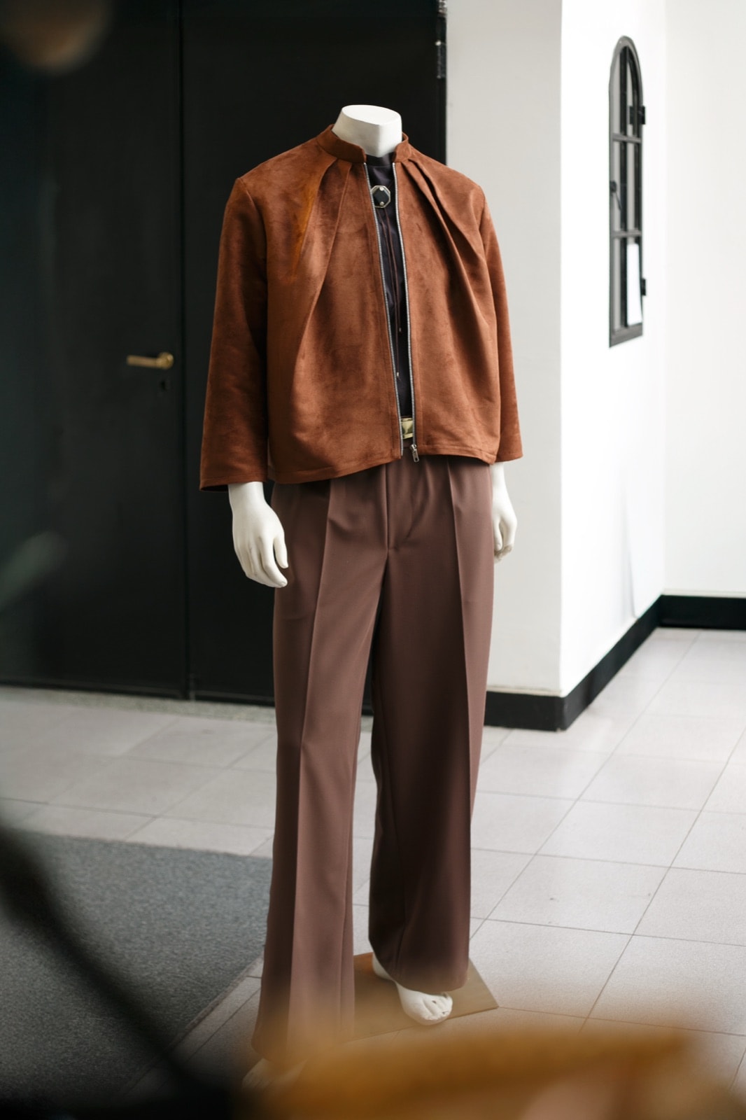 Chaqueta de gamuza caramelo sobre pantalón marrón, vista delantera completa sobre maniquí en la entrada del showroom.