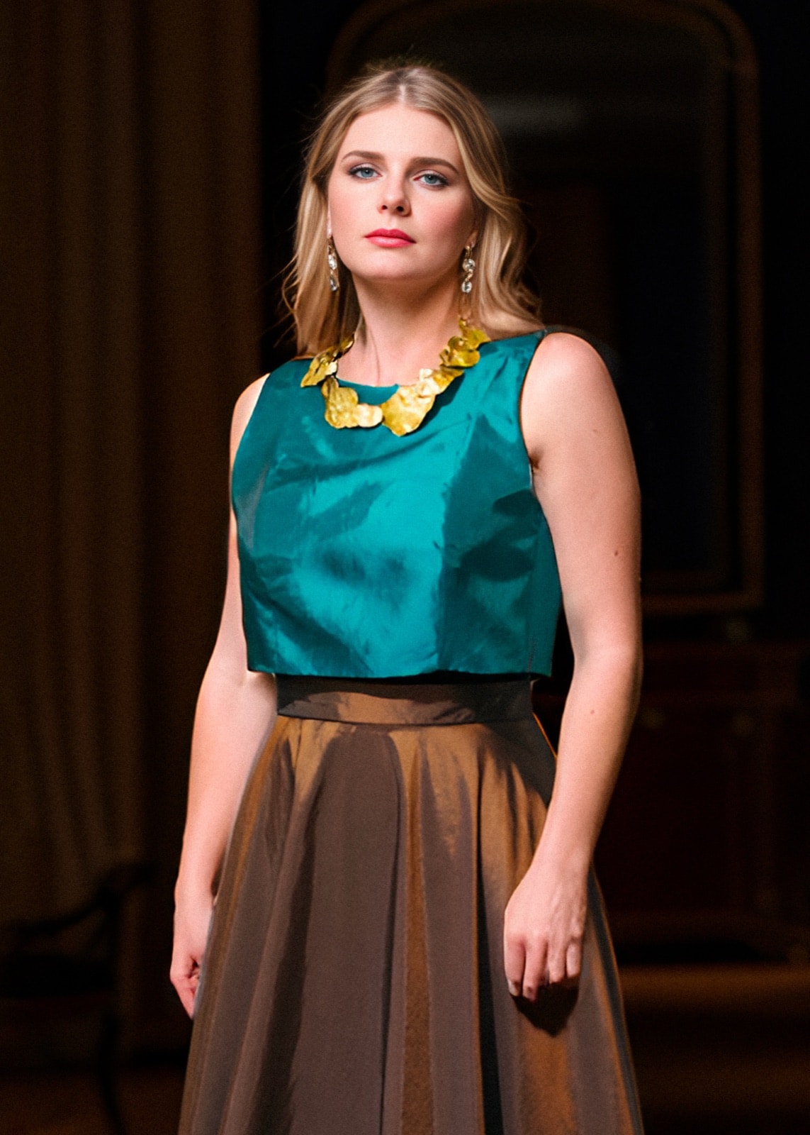 Croptop de tafetán petróleo de Atelier Molina sobre modelo, combinado con falda marrón y collar dorado en un salón oscuro.