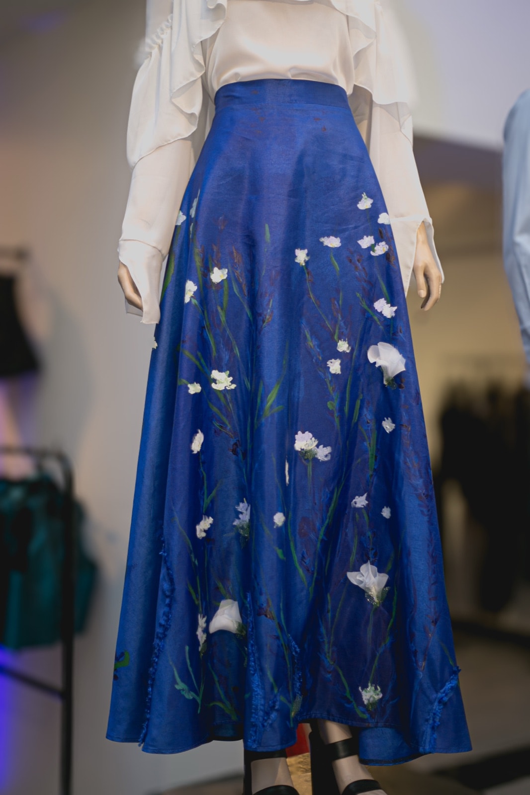 Falda de tafetán azul con jardín pintado y bordado sobre maniquí, plano cercano centrado en la caída del vuelo delantero.