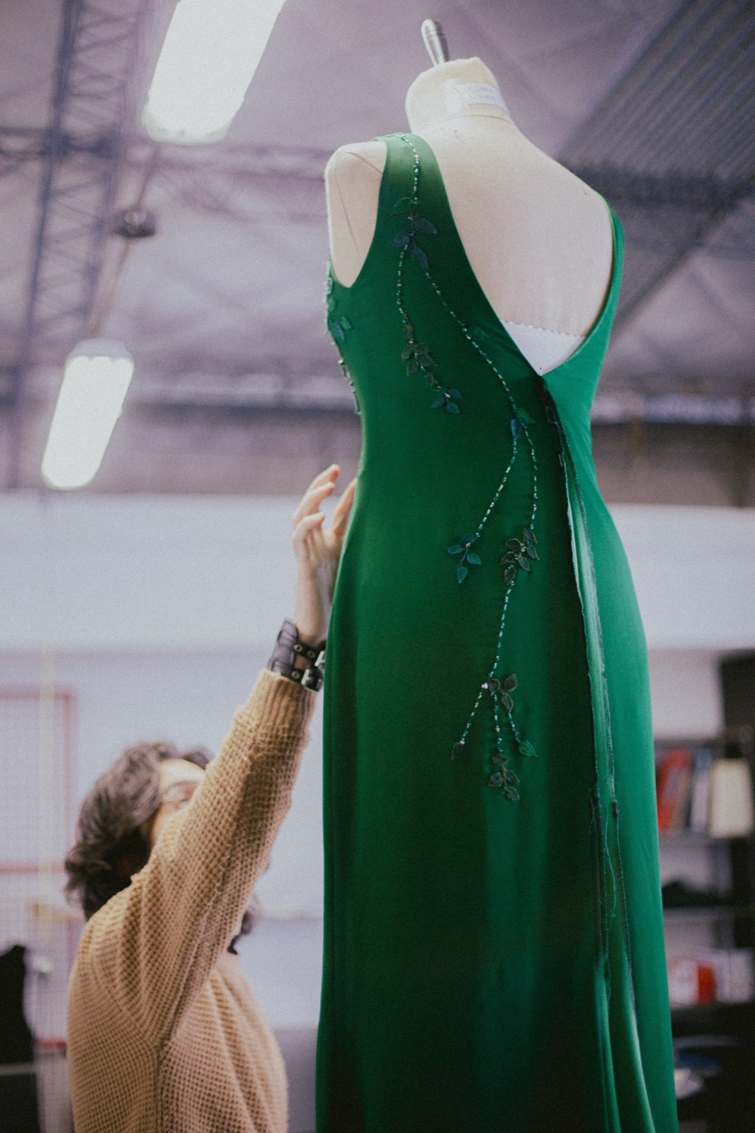 Ajuste en el taller del vestido verde sobre maniquí, con la modista sosteniendo la tela para revisar la caída de la falda por detrás.