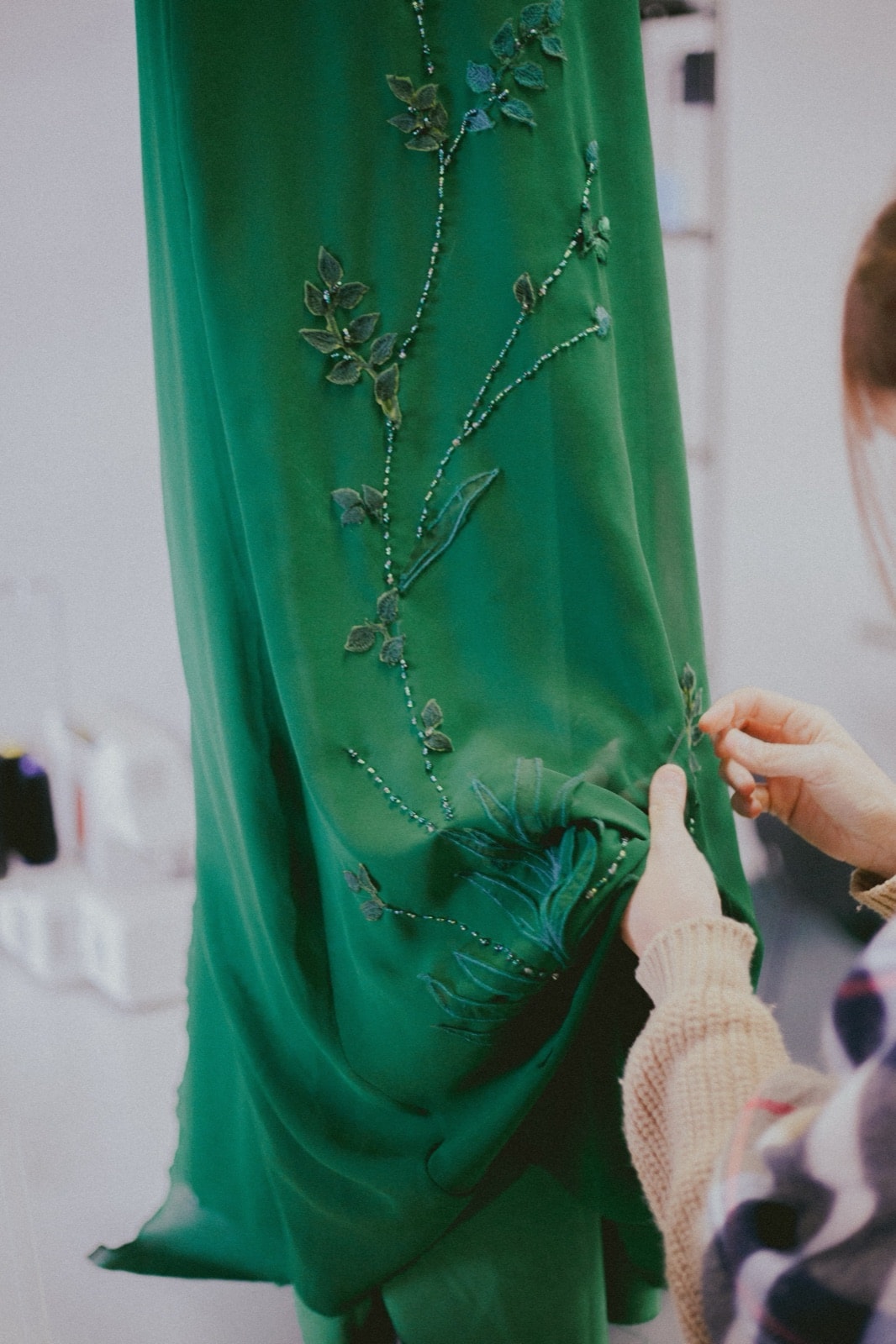 Detalle del bordado en la falda del vestido verde, con una mano sosteniendo el pliegue de gasa para mostrar las hojas bordadas.