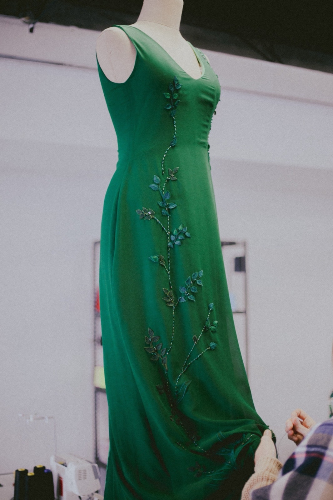 Vestido largo verde sobre maniquí en el taller, vista de perfil que muestra el escote en V y el bordado floral a lo largo del cuerpo.