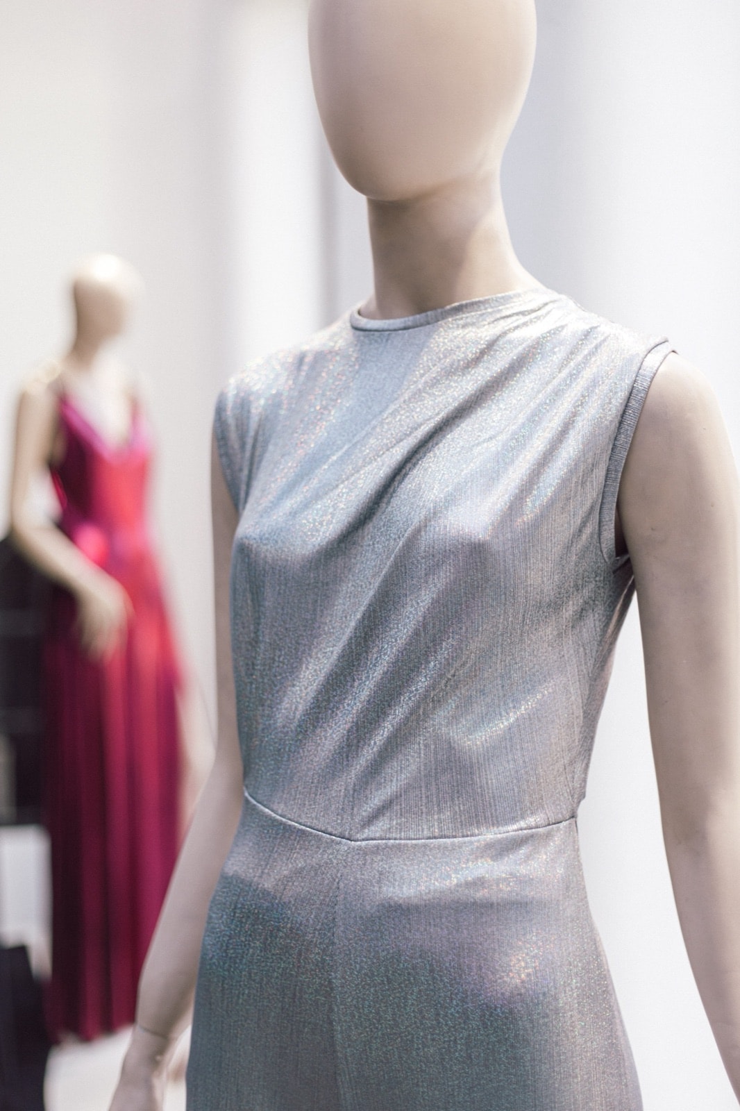 Detalle del torso del mono plateado holográfico sobre maniquí, con un vestido rosa desenfocado de fondo en el showroom.