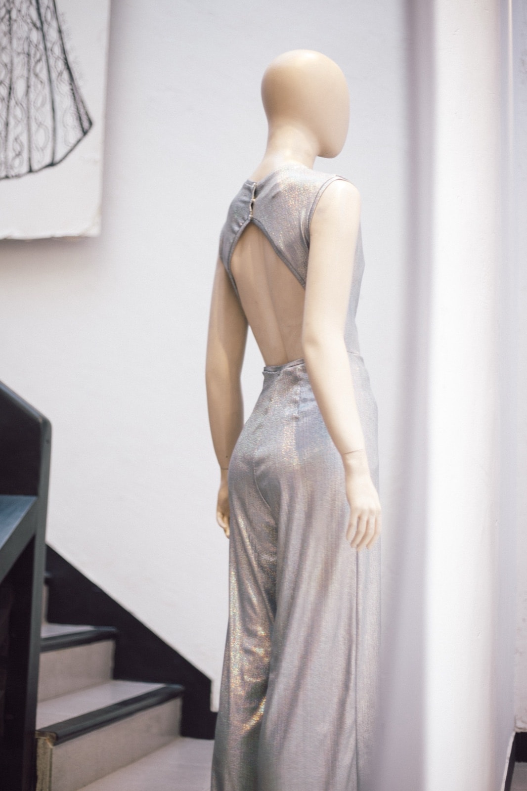 Vista trasera completa del mono plateado holográfico sobre maniquí, con el recorte abierto en la espalda junto a una escalera.