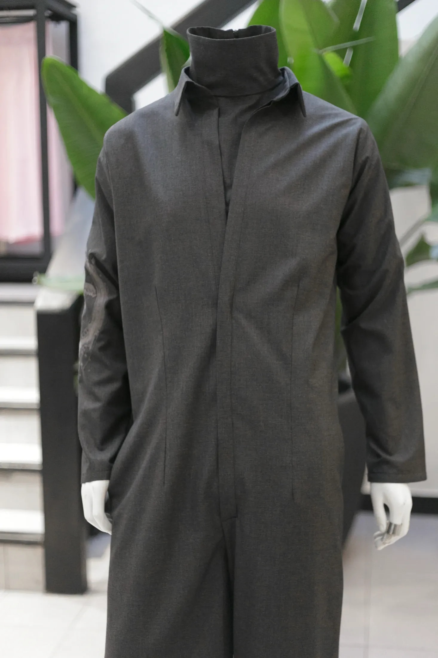 Vista del torso del jumpsuit sastrero gris sobre maniquí, con el bordado del brazo asomando junto a una escalera del showroom.