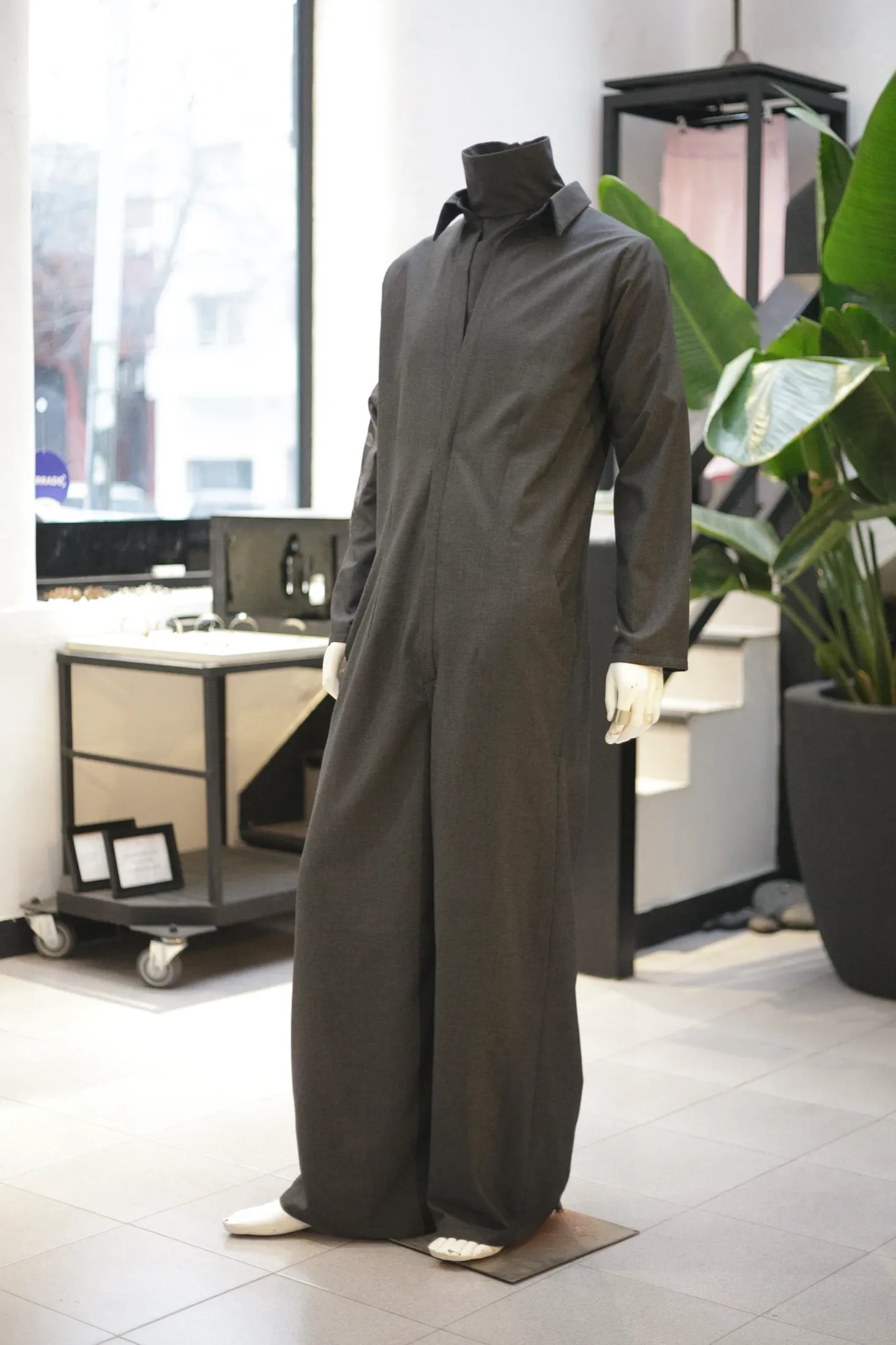 Jumpsuit sastrero gris sobre maniquí de cuerpo entero en el showroom, con plantas y vitrinas de accesorios de fondo.