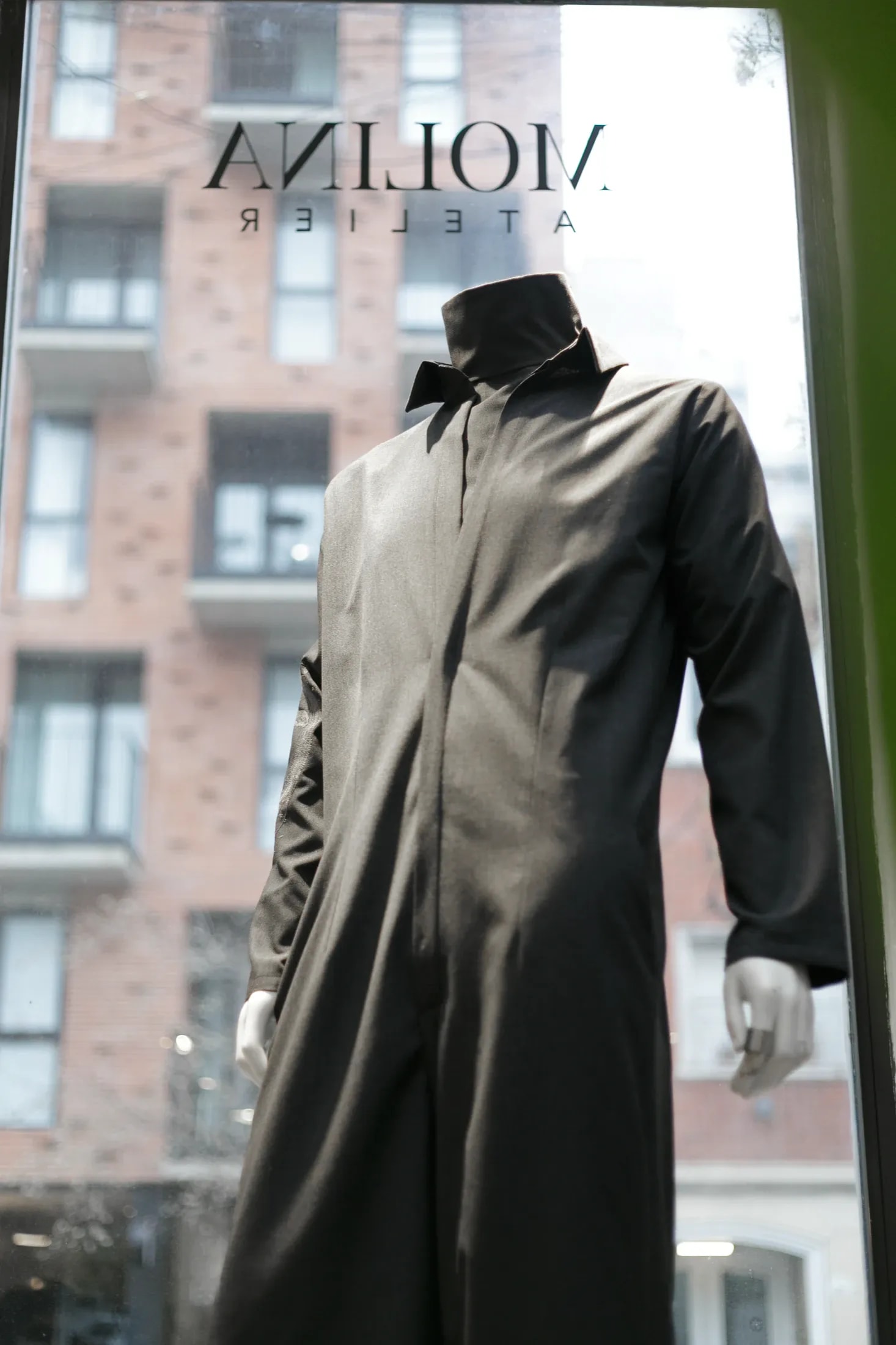 Jumpsuit sastrero gris sobre maniquí en la vidriera del showroom de Atelier Molina, con el edificio de enfrente reflejado en el vidrio.