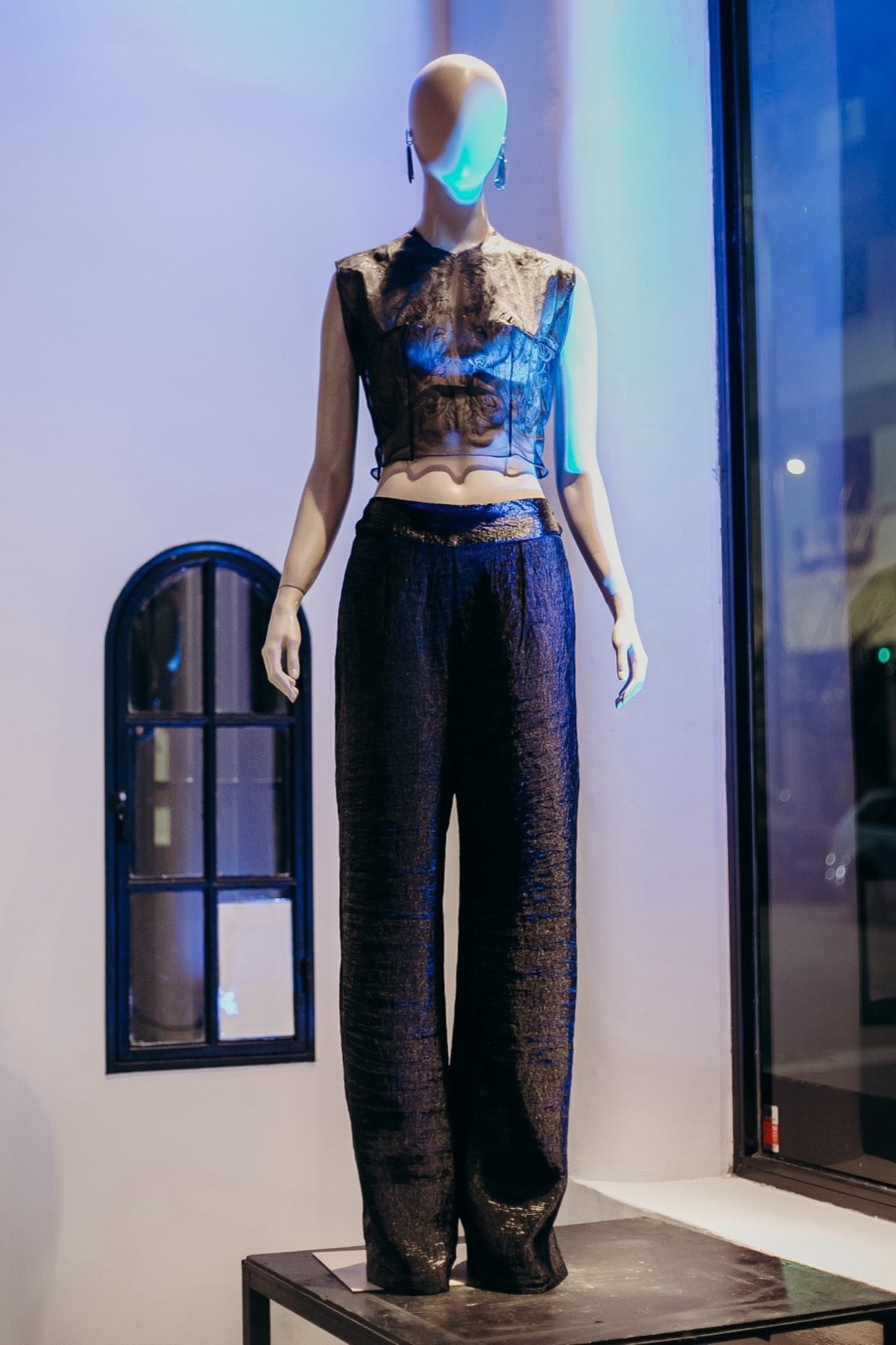 Pantalón ancho texturado negro sobre maniquí con top de encaje transparente, vista frontal completa junto a la ventana.
