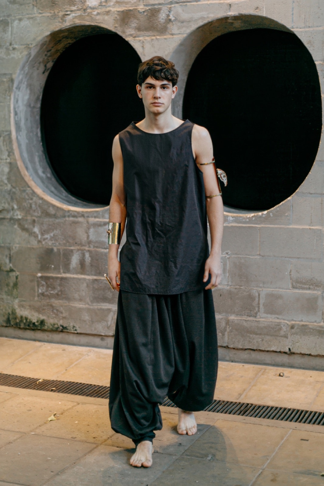 Pantalón chiripá gris oscuro de Atelier Molina sobre modelo de cuerpo entero, de pie frente a un muro con dos aberturas circulares.