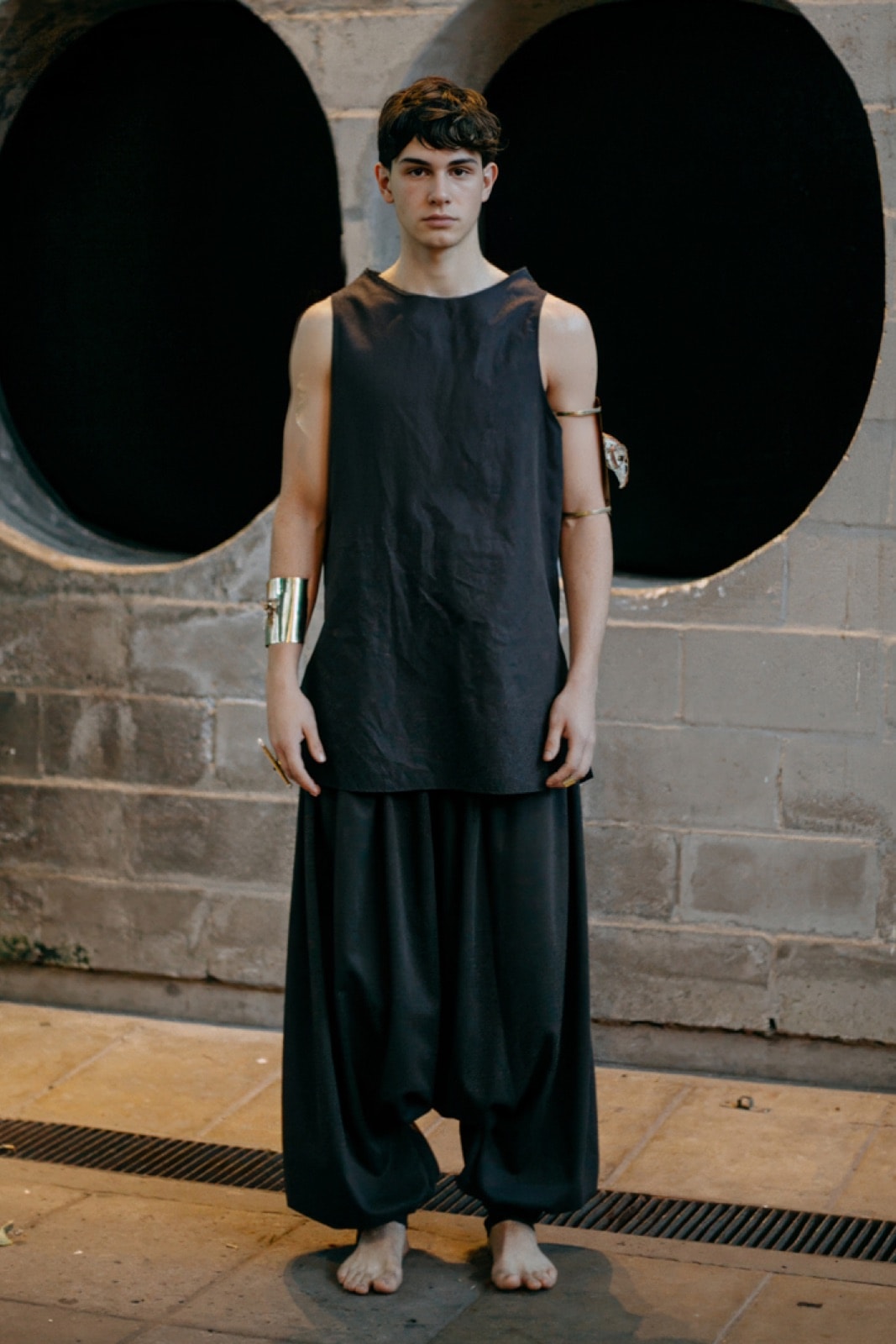 Pantalón chiripá gris oscuro de Atelier Molina sobre modelo, otro encuadre de cuerpo entero frente al mismo muro de piedra.