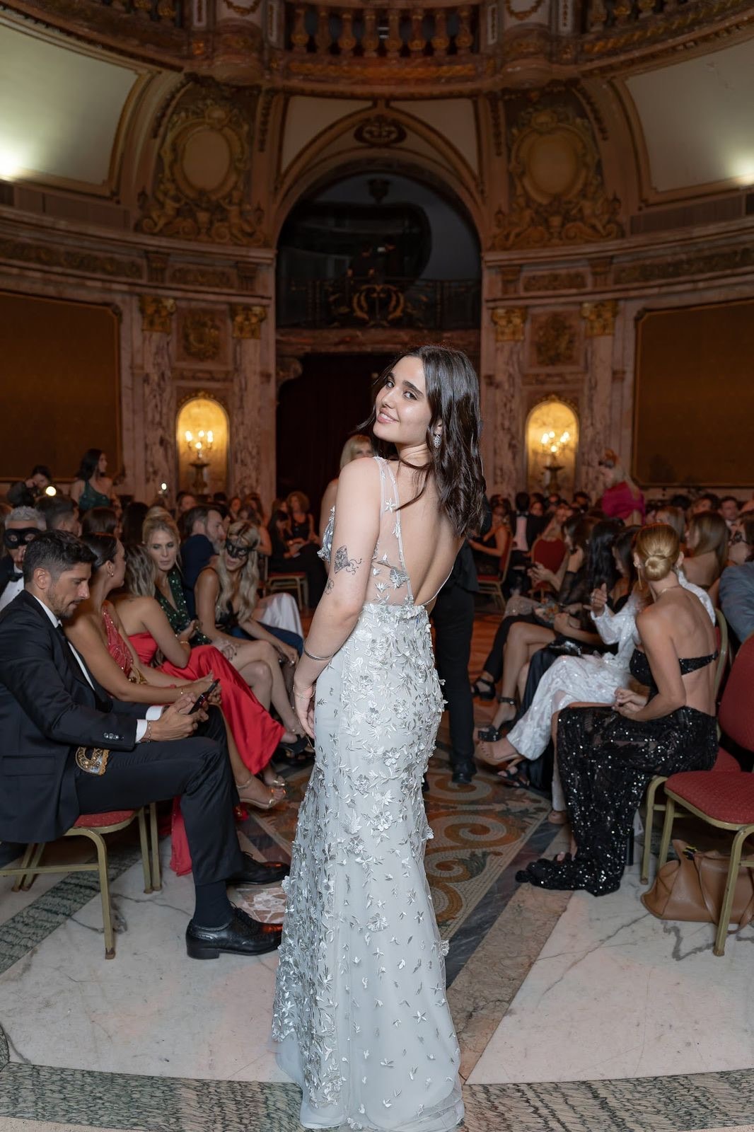 Espalda del vestido de tul plateado bordado de Atelier Molina, sobre modelo en un salón de gala con público sentado alrededor.