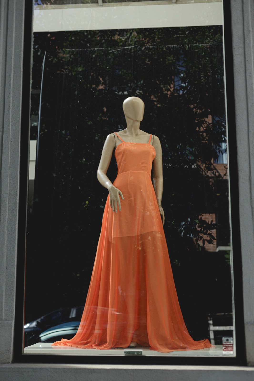Sunset Glow Dress naranja de Atelier Molina sobre maniquí en una vidriera, con el follaje de la calle reflejado en el vidrio.
