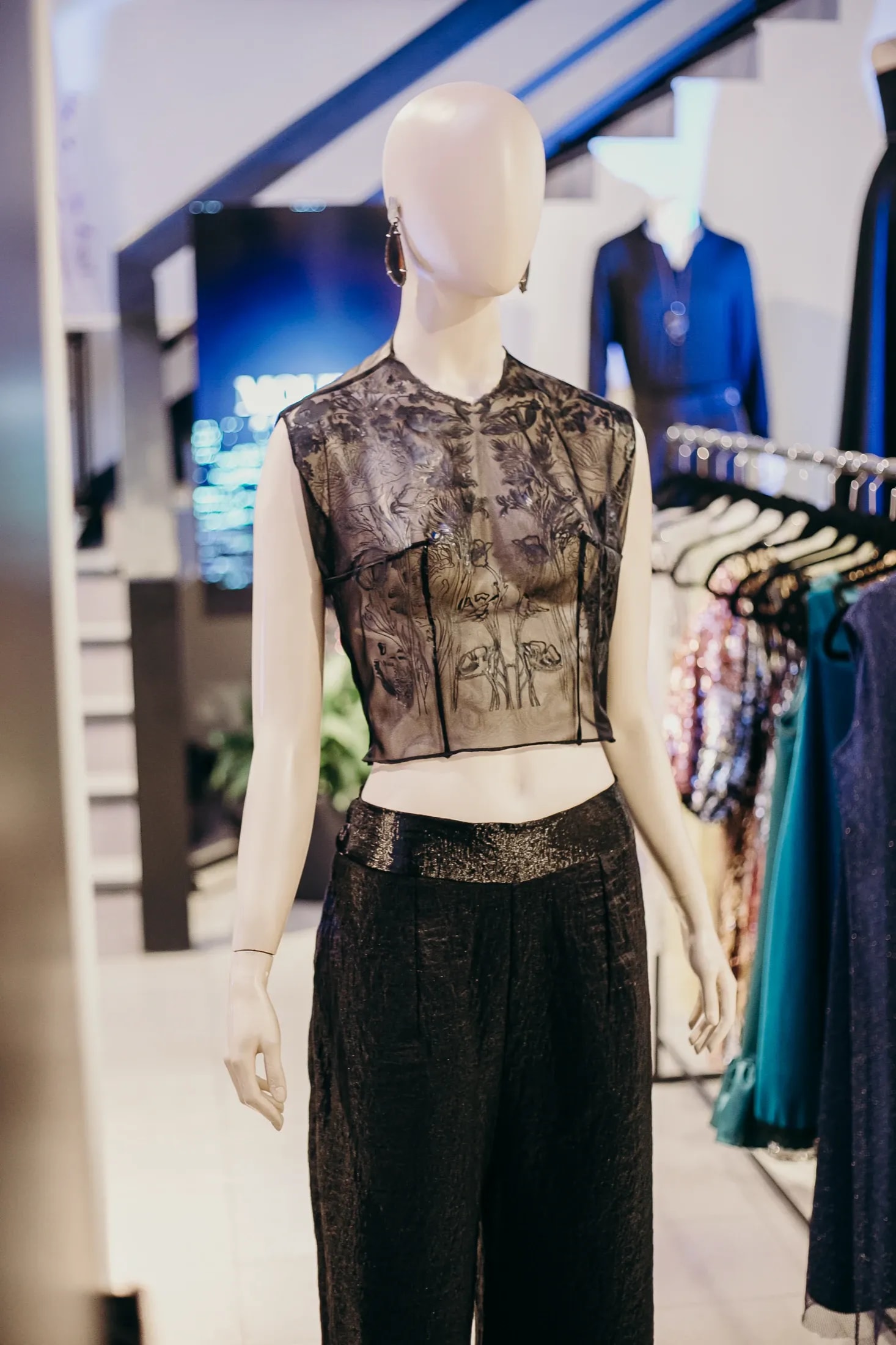 Top negro transparente con estampado de flores y mariposas sobre maniquí en el showroom, combinado con pantalón negro.