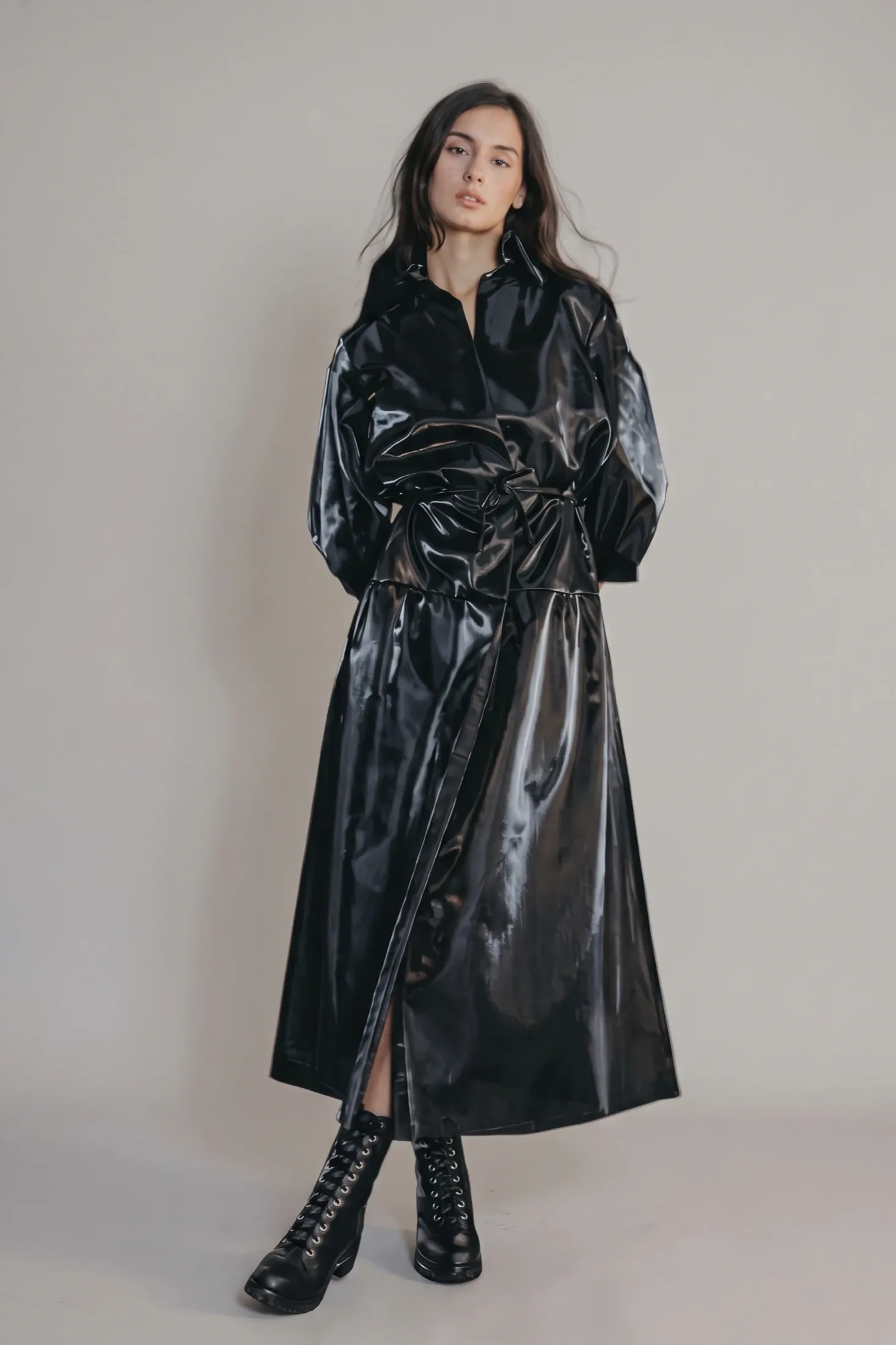 Trench largo negro de acabado brillante de Atelier Molina, Buenos Aires, de frente con cintura ajustada, falda amplia y botas negras de cordones.