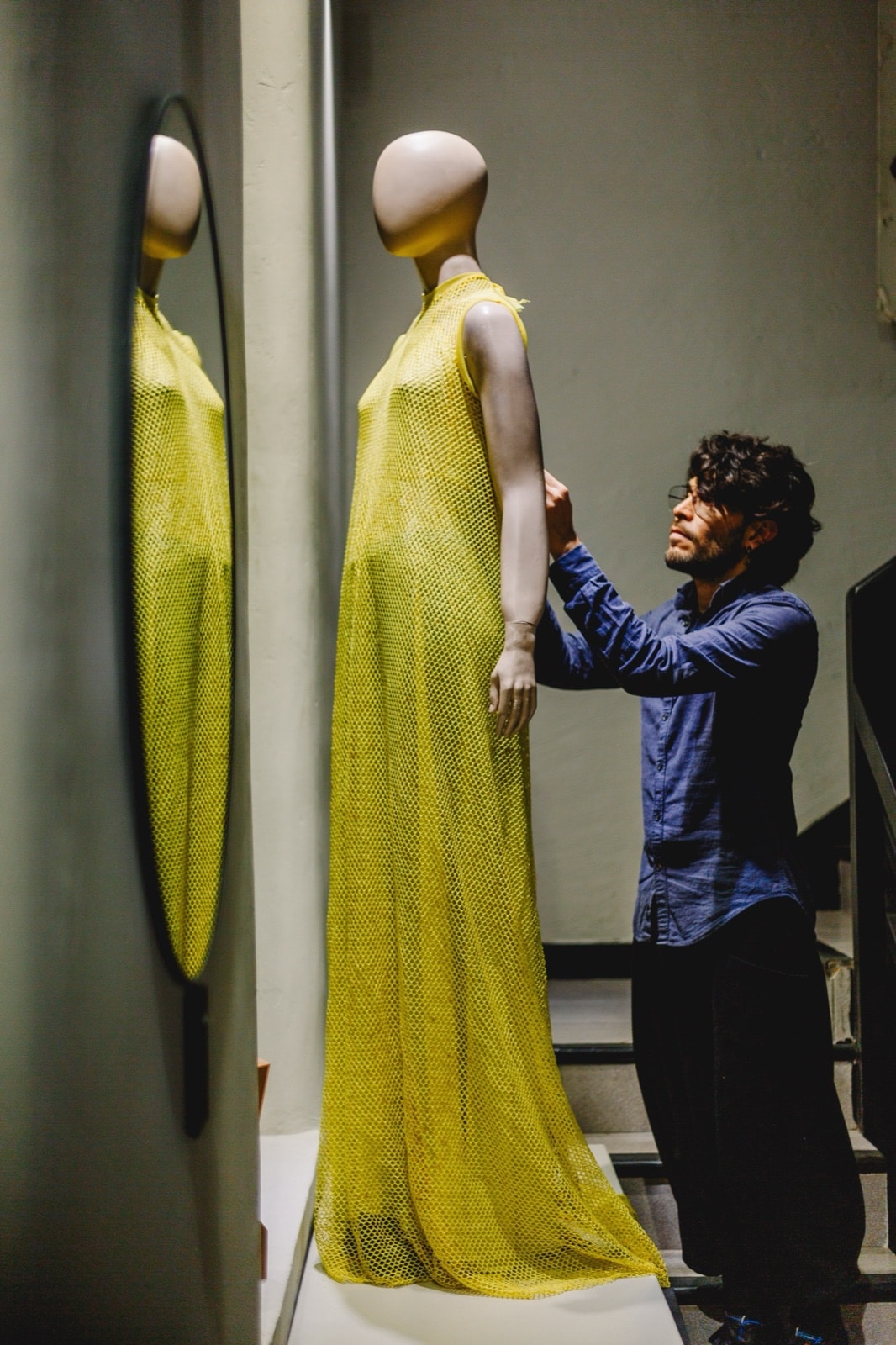Ajuste de la túnica de red amarilla sobre maniquí en la escalera del taller, con un espejo que refleja la caída de la tela.