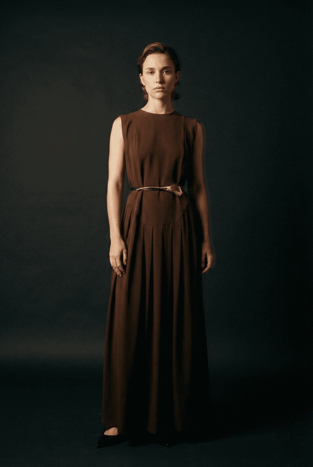 Vestido Chocolate con Tablas y Cinturón Cala sobre modelo, vista frontal completa con iluminación tenue sobre fondo oscuro.
