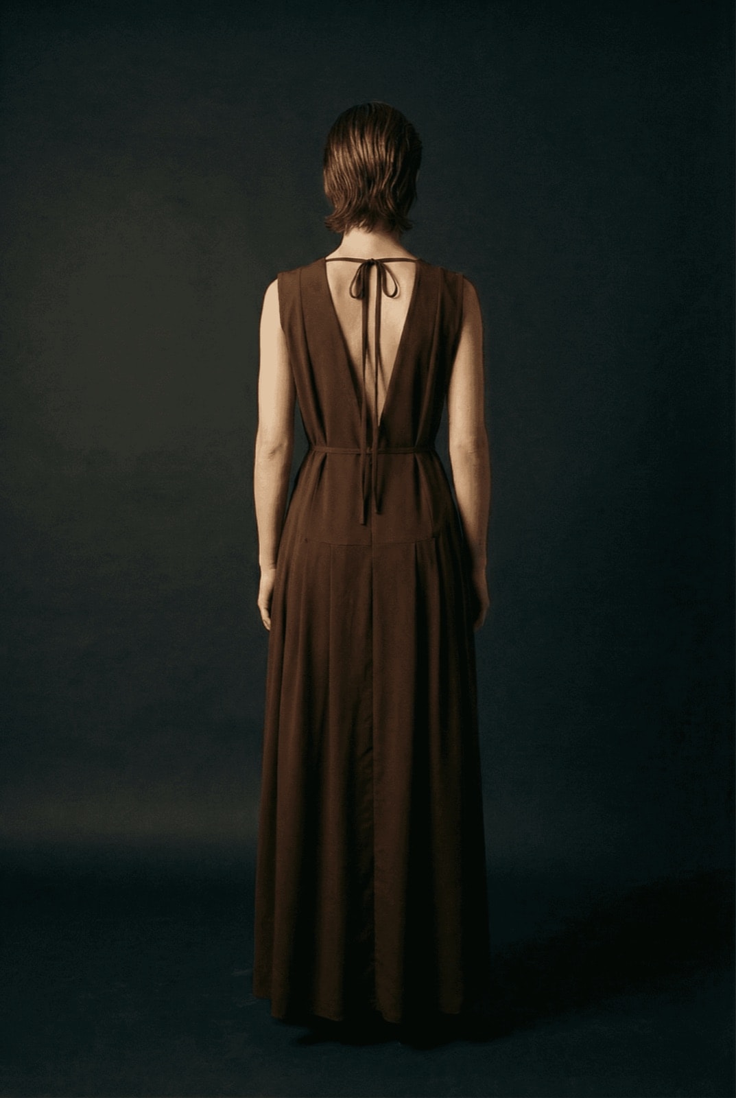 Vestido Chocolate con Tablas y Cinturón Cala sobre modelo, vista de espalda con el escote profundo y el lazo del cierre.