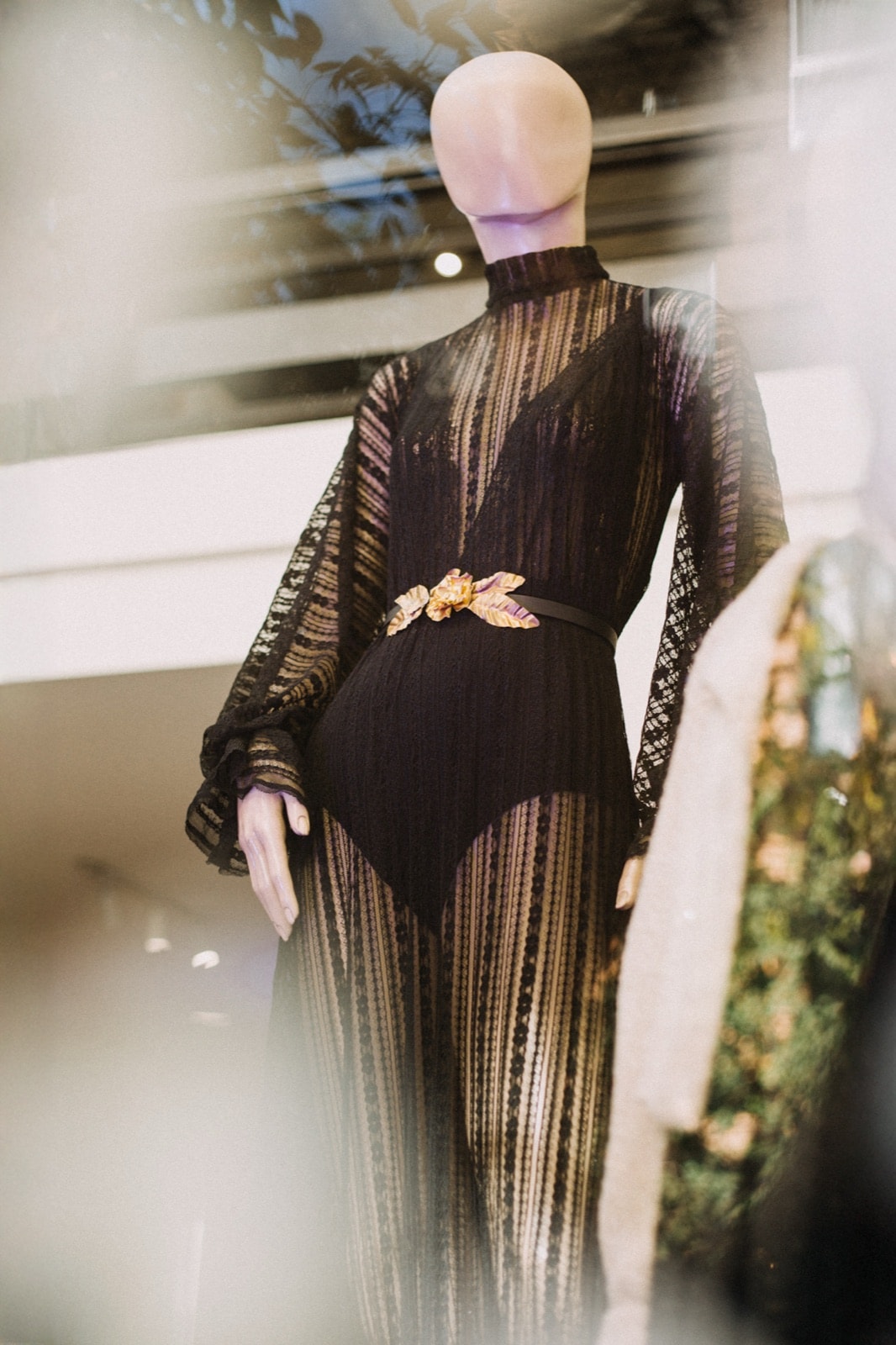 Vestido de encaje vertical elastizado negro sobre maniquí en vidriera, primer plano del cinturón con aplique floral dorado.