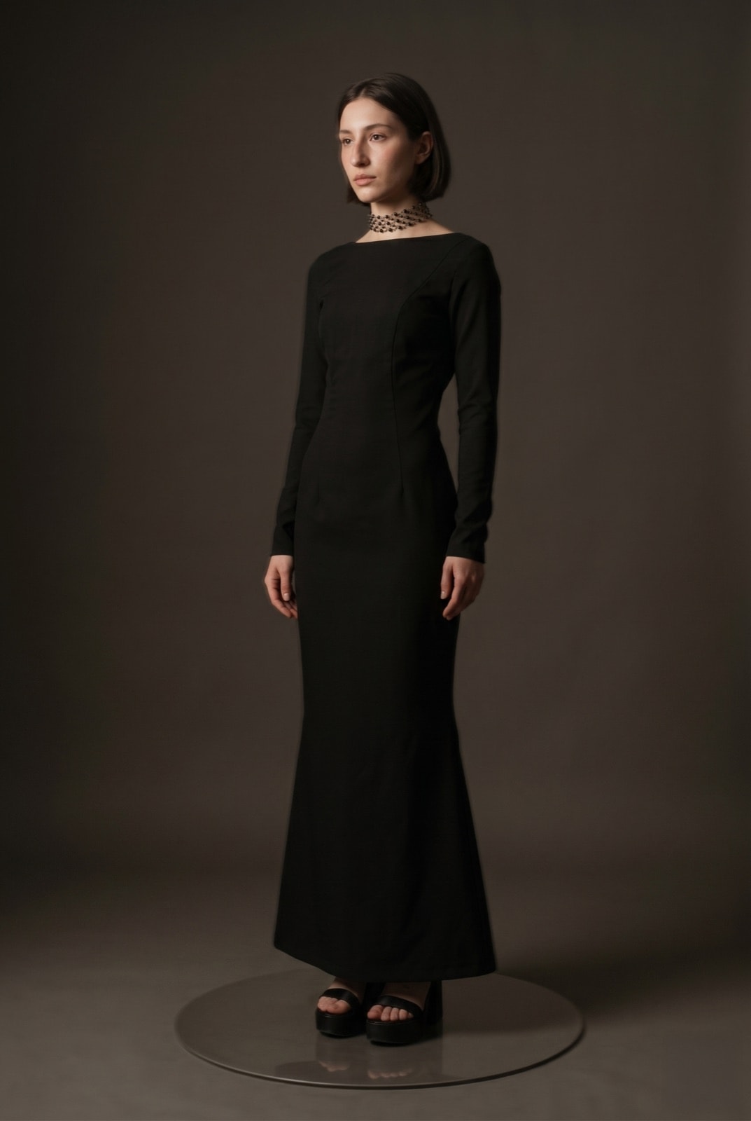 Vestido largo negro de gabardina de Atelier Molina, vista de tres cuartos sobre modelo, fondo marrón oscuro de estudio.