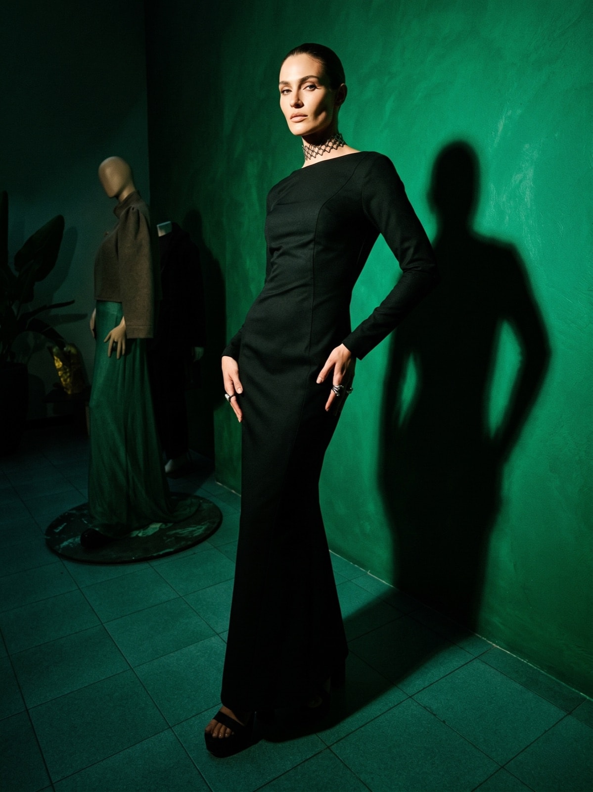 Vestido largo negro de gabardina de Atelier Molina, sobre modelo de pie junto a un maniquí, fondo verde del showroom.