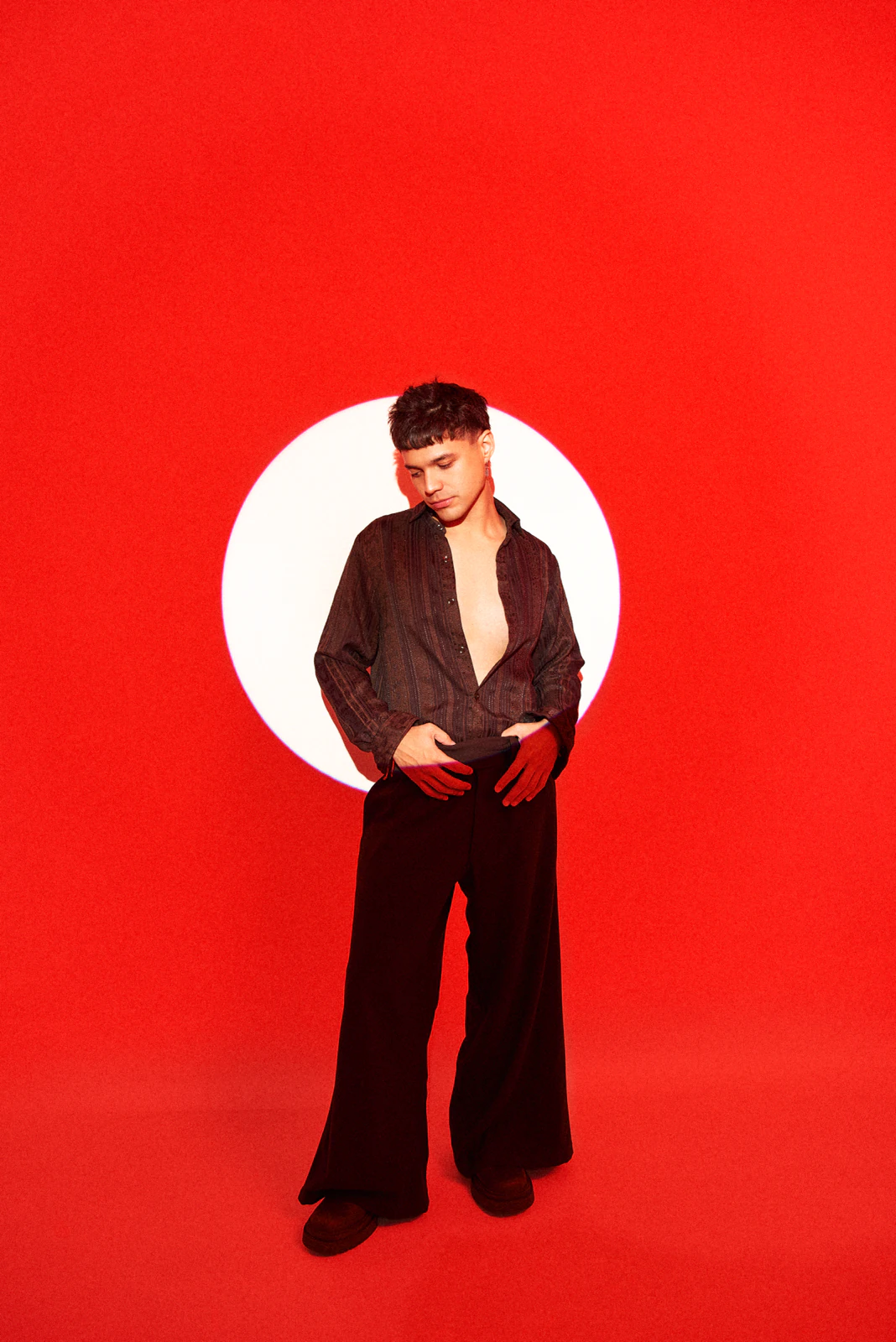Alan Lez against a red backdrop in a sheer shirt and black palazzo trousers by Atelier Molina