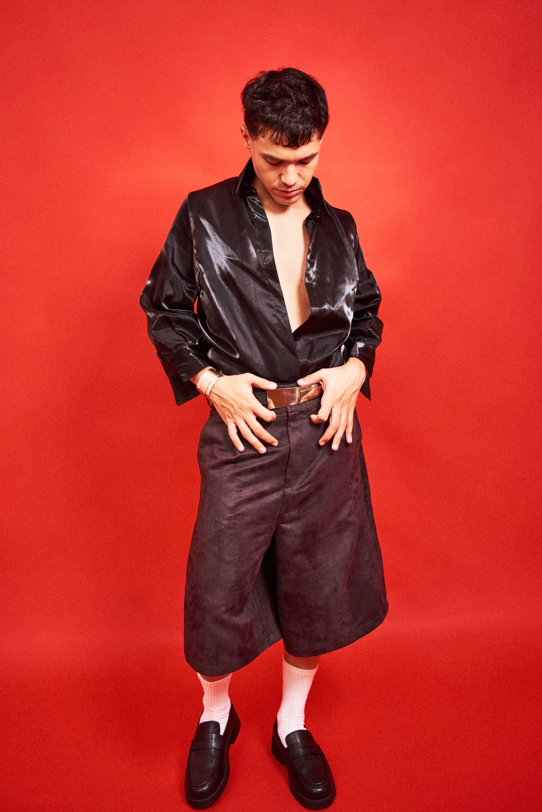 Alan Lez in a black satin shirt and tailored bermuda shorts by Atelier Molina against a red backdrop
