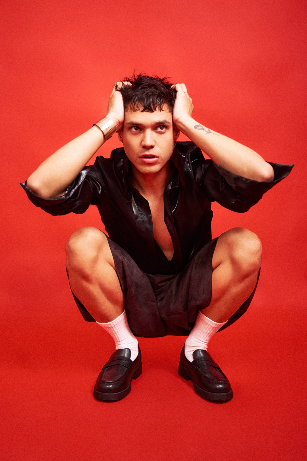Alan Lez crouching against a red backdrop in a black shirt and bermuda shorts by Atelier Molina