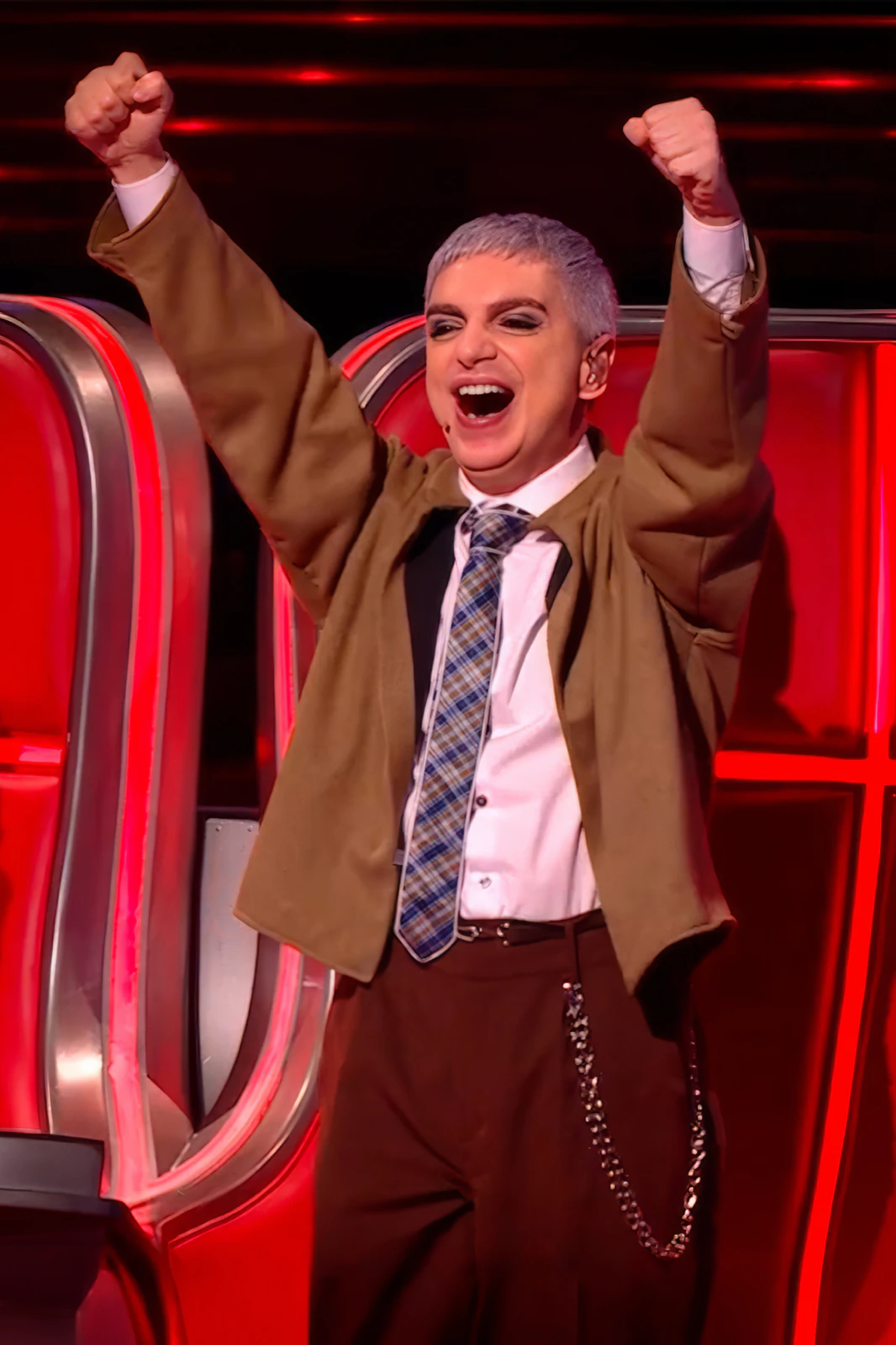 Ale Sergi raising both arms on the La Voz Argentina set, in a camel jacket and plaid tie by Atelier Molina